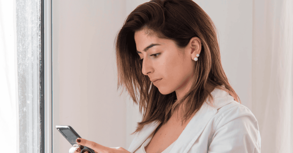 A young woman looks down at her smartphone with a neutral, focused expression, signaling a shift in perspective that leads to a new kind of emotional distance.