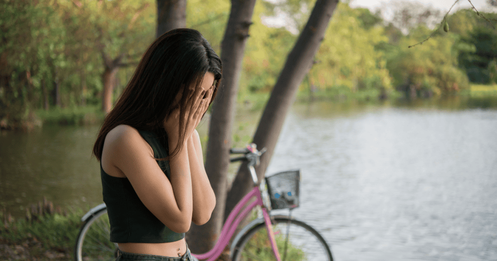 A woman stands by a quiet lake with her face buried in her hands, depicting the profound disorientation that follows the collapse of a central life pillar.