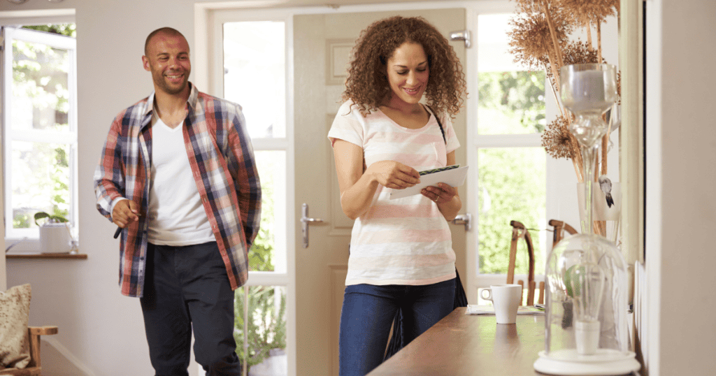 A woman smilingly reviews letters at a wooden counter while a man walks into the home behind her, portraying a scene of domestic normalcy that underscores a carefully planned personal pivot.