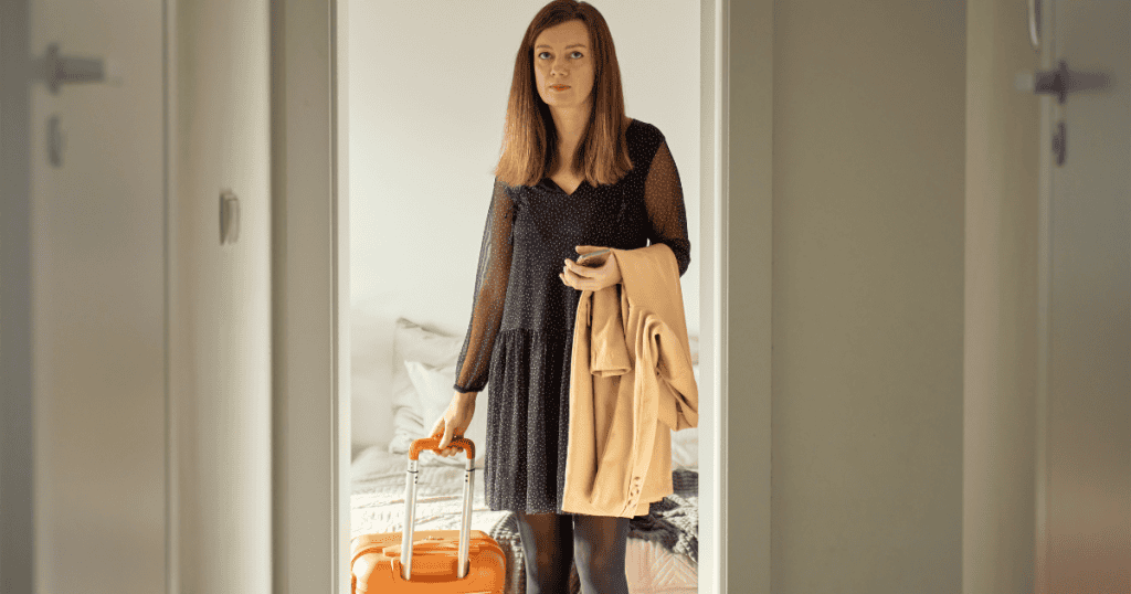 A woman stands firmly in a doorway holding a suitcase handle and a trench coat, capturing the precise, understated moment an intentional departure is set into motion.