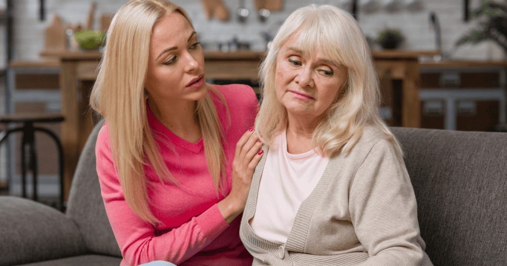 A younger woman places a hand on the shoulder of a somber older woman on a sofa, capturing a moment governed by an unspoken social contract that neither is willing to break.
