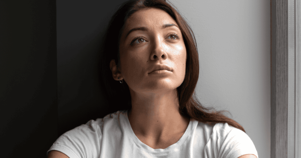 A woman in a white t-shirt leans against a dark wall and stares upward with a somber, clarity-seeking look, signaling a sudden internal shift away from a long-held illusion.