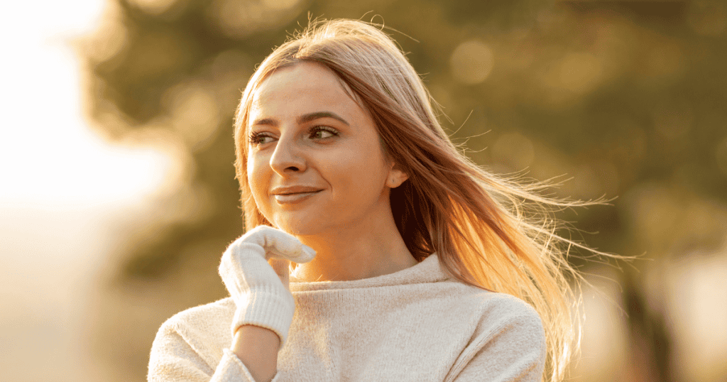 A young woman in a white sweater smiles softly while looking out into the warm evening light, illustrating the serene confidence that surfaces when one decides to step away from an uneven pursuit.