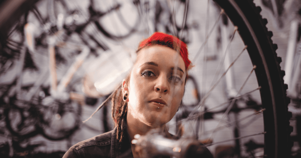 A woman with bright red hair and facial piercings looks directly through the spokes of a bicycle wheel with a steady, solemn gaze, representing the intricate structural web that only becomes visible after stepping back from it.