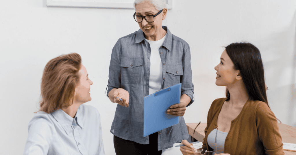 An older woman in a denim shirt holds a clipboard and smiles at two younger colleagues in an office setting, highlighting the polished exterior presented to the world versus the reality behind closed doors.