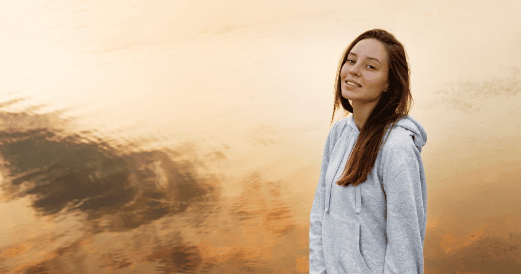 A woman in a gray hoodie looks toward the camera with a slight, knowing smile against a wide expanse of still water at dusk, capturing a moment where an empty silence is recognized as a deliberate tactic.