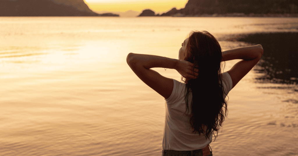 A woman stands with her back to the camera facing a vast, golden sunset over water, her relaxed posture reflecting a quiet acceptance of a reality that has become her only horizon.