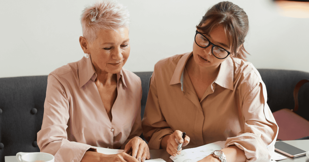 Two women sit closely together reviewing documents with focused expressions, capturing a subtle moment of collaboration where the balance of power quietly begins to tilt.