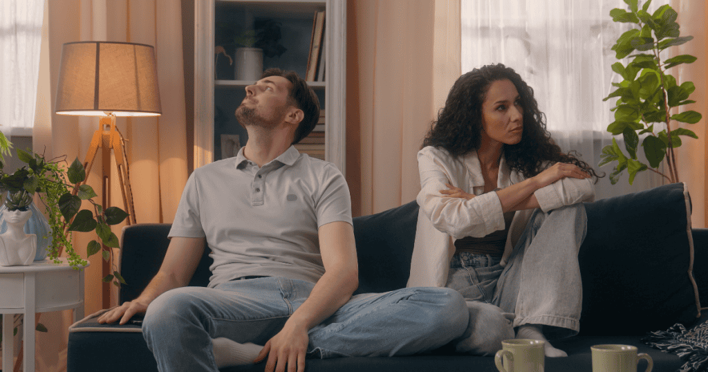A man stares blankly upward while a woman sits next to him on a sofa with her arms tightly crossed and a tense expression, showcasing the heavy, artificial distance created by an intentional refusal to engage.
