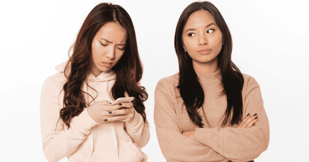 One young woman looks down anxiously at her smartphone screen while her companion stands with crossed arms and an expectant, uneasy sidelong glance, portraying the subtle mental strain of waiting on someone else's terms.