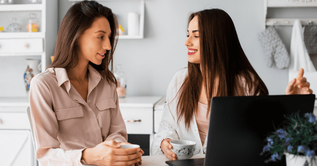 Two young women sit at a kitchen table smiling over coffee and a laptop, portraying a supportive exchange that carries an unspoken expectation of future repayment.