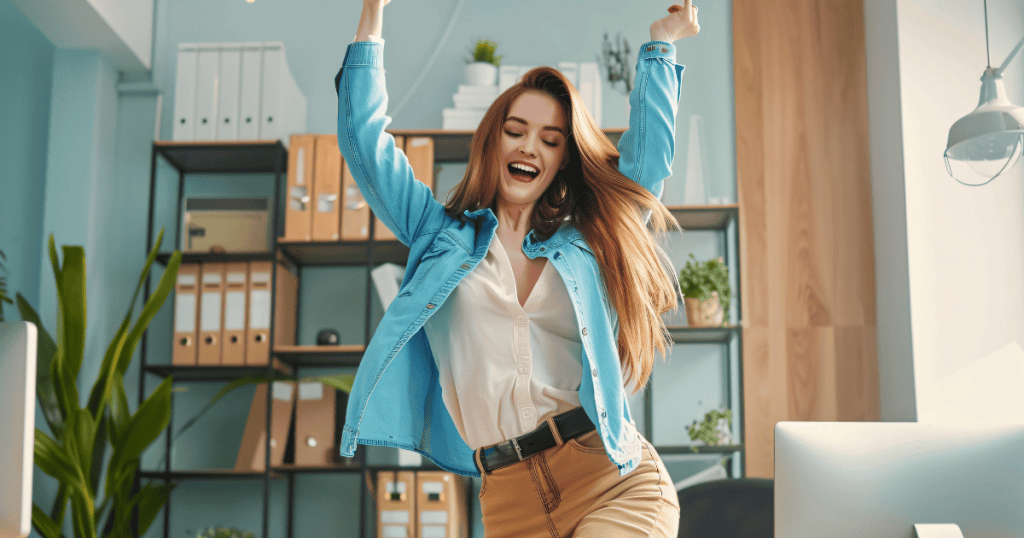 A woman in a blue shirt dances with her arms raised high in a bright office, celebrating the liberation of remaining connected to her own joy regardless of the setting.