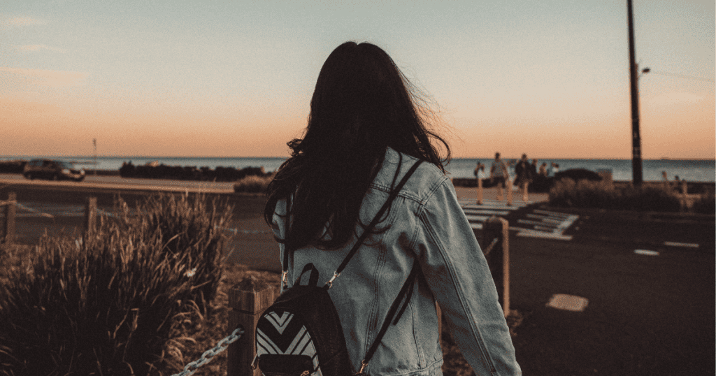 A woman wearing a denim jacket and a backpack walks away down a coastal road toward a fading sunset, symbolizing the deliberate choice to keep moving forward into a new dawn.