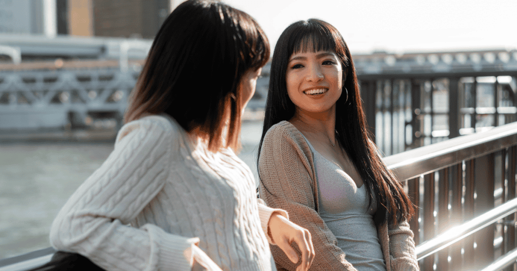 Two women talk near a riverside railing while one smiles warmly and leans in, capturing a disarming and seemingly pleasant interaction that makes it easy to overlook underlying intentions.