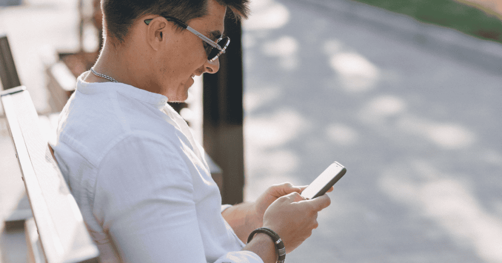 A young man wearing sunglasses sits on a park bench and smiles subtly while typing on his phone, showcasing the casual, low-stakes digital reach-out used to re-establish a connection.