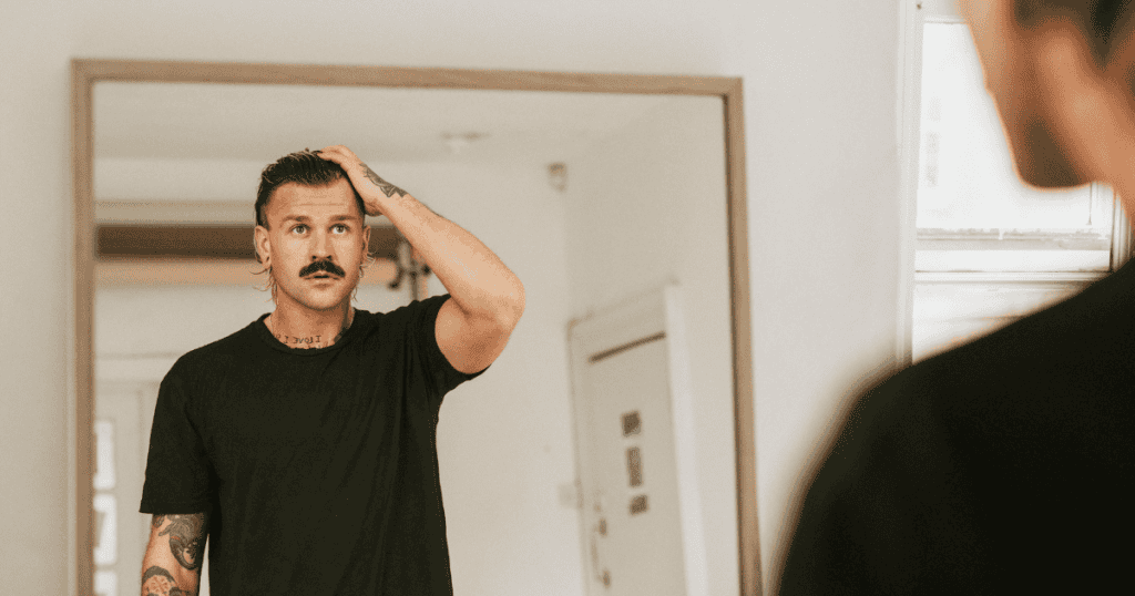 A man examines his reflection in a mirror while adjusting his hair, focused on the preservation of a curated facade.