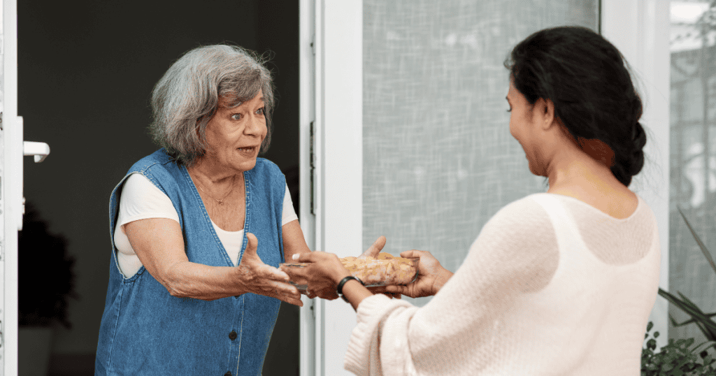 An older woman stands in a doorway excitedly handing a dish of food to another woman, illustrating how an overt act of generosity can be fueled by a deep-seated need for validation.