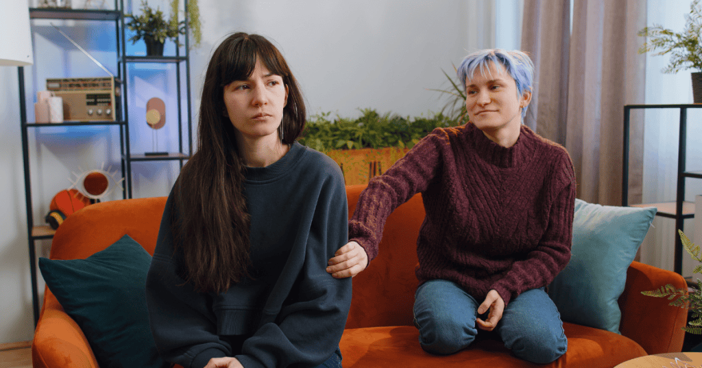 A woman with a somber expression sits on a sofa while another individual with short lavender hair kneels behind her and casually rests a hand on her arm, depicting a sudden, familiar approach that completely ignores past friction.