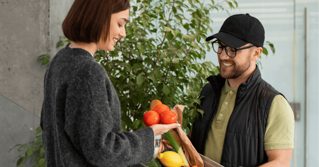 A man in a cap smiles warmly while handing a bag of fresh produce to a woman, showcasing the disarming nature of a helpful, friendly interaction that builds immediate rapport.