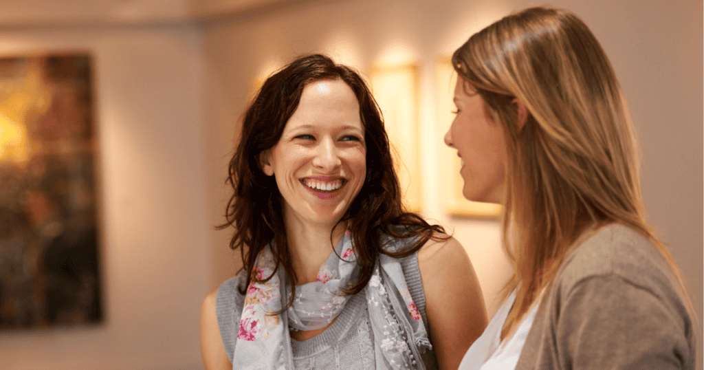 A woman with dark curly hair laughs radiantly during a bright conversation in an art gallery, embodying the exact moment personal joy returns, which often serves as a beacon for an unexpected reappearance.