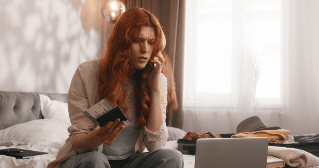 A red-haired woman on a bed looks distressed while holding tickets and talking on her phone next to an open suitcase, illustrating a sudden, manufactured crisis that preys on her natural urge to help.