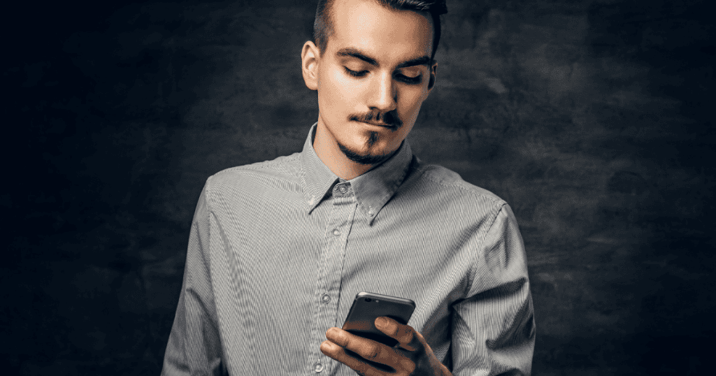 A young man with a neat goatee looks down quietly at his mobile phone against a dark background, capturing a solitary moment of withholding attention to create an unresolved space.