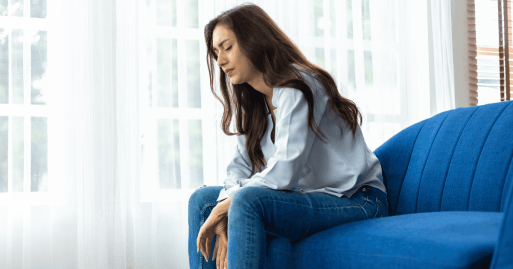 A woman sits on the edge of a bright blue sofa with her head lowered and eyes closed in deep exhaustion, embodying the heavy invisible ties that make breaking free feel completely draining.