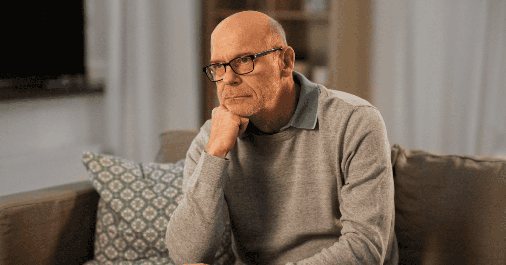 A man sits alone on a couch with a heavy, contemplative expression, grappling with a pain that feels disproportionate to the visible facts.