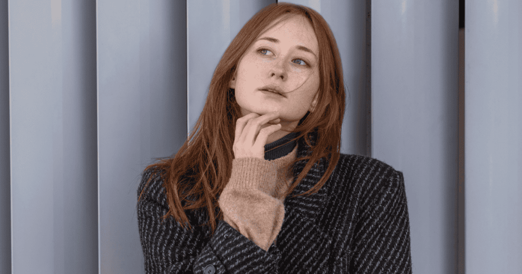 A young woman with red hair rests her chin on her hand and gazes upward with a contemplative look, reflecting the internal confusion that arises when reality doesn't match a perceived kindness.