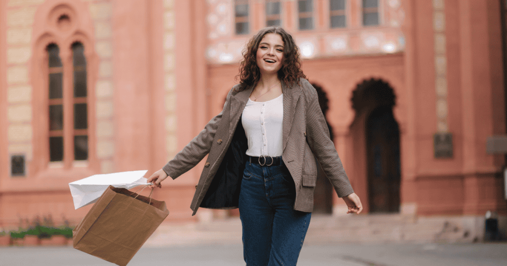 A young woman with curly hair smiles radiantly while walking outside an ornate brick building with shopping bags, embodying a profound sense of personal elevation and liberty.