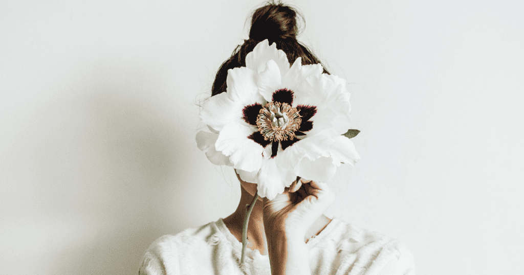 A person holds a large white flower directly in front of their face, symbolizing the temporary obscuring of one's true identity during a period of intense focus on someone else.