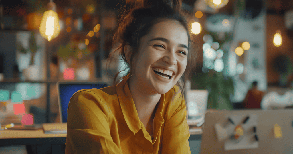 A woman shares a genuine, wide-mouthed laugh in a warm office environment, embodying a sense of ease that comes from abandoning a curated facade.
