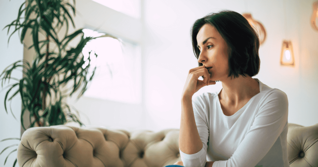 A woman sits thoughtfully on a tufted beige sofa with her hand resting against her chin, representing the quiet, analytical focus required to secure one's own future security.