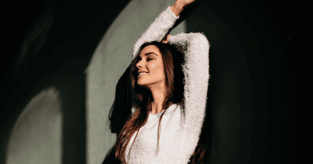 A woman leans against a wall with her arms raised and eyes closed in the sunlight, embracing a newfound sense of release after the silence.