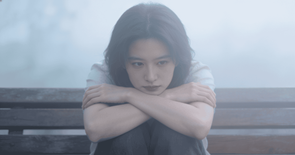 A young woman sits curled up on a wooden bench while surrounded by a thick, heavy mist, visually representing the disorienting sensation of brain fog.