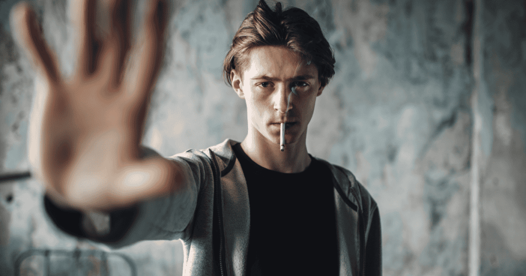 A young man holding a smoking cigarette holds his hand out to directly block the camera lens, signaling a sharp boundary to stop you from questioning his actions.