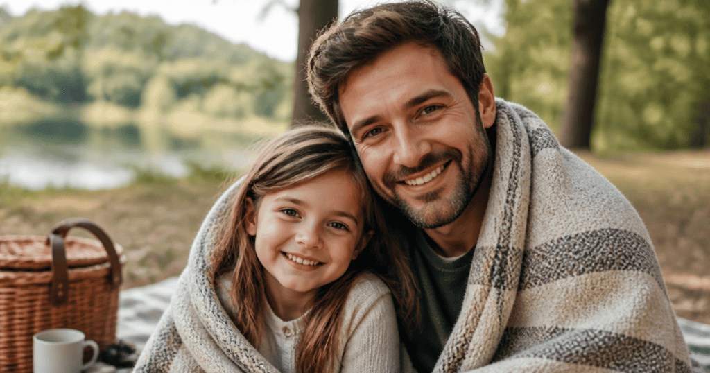 A smiling man and his young daughter are wrapped together under a plaid blanket during an outdoor picnic, subtly hinting at the early bonding dynamics behind 7 childhood patterns that keep pulling you toward narcissists.