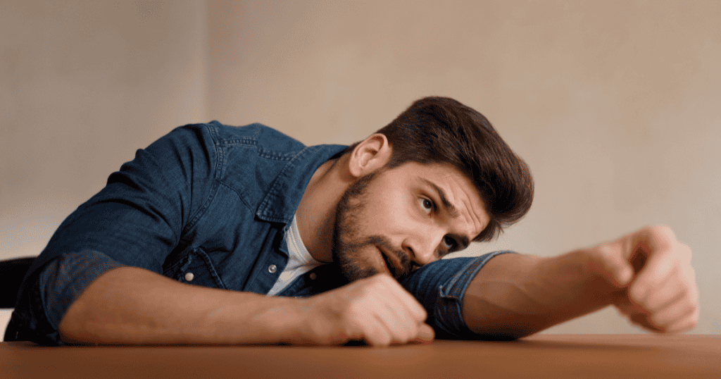 A man slumped lazily over a table with a defiant look, depicting how a narcissist deliberately feigns helplessness and incompetence to avoid responsibility and shift the burden onto others.