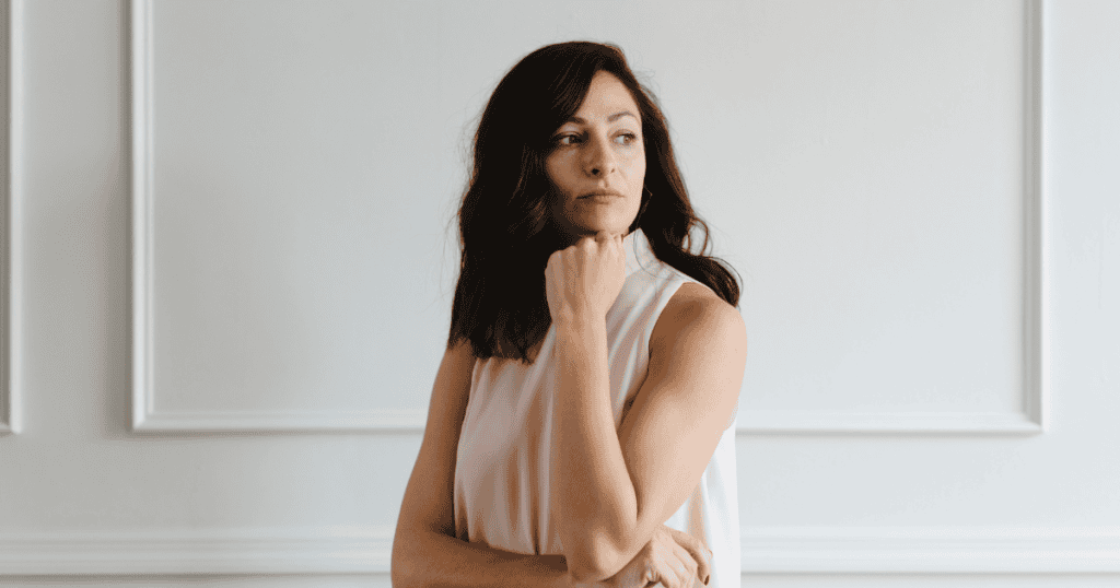 A pensive woman in a light sleeveless top rests her chin on her hand while looking off to the side, quietly processing how someone completely ruined her mood.