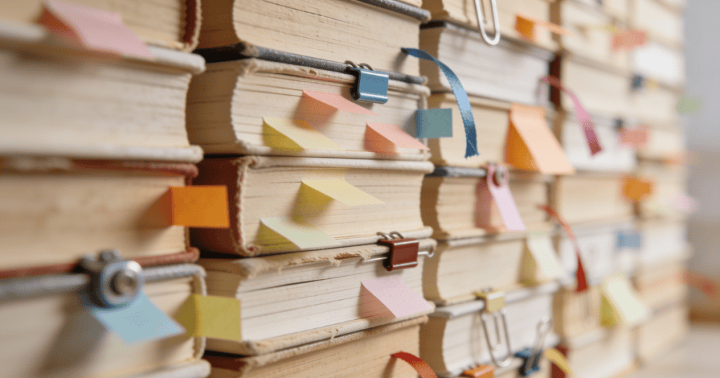 A close-up view shows multiple stacks of old, weathered textbooks heavily tagged with colorful sticky notes and paperclips, representing a deep and rigorous study into the systemic mechanics behind long-lasting behavioral cycles.