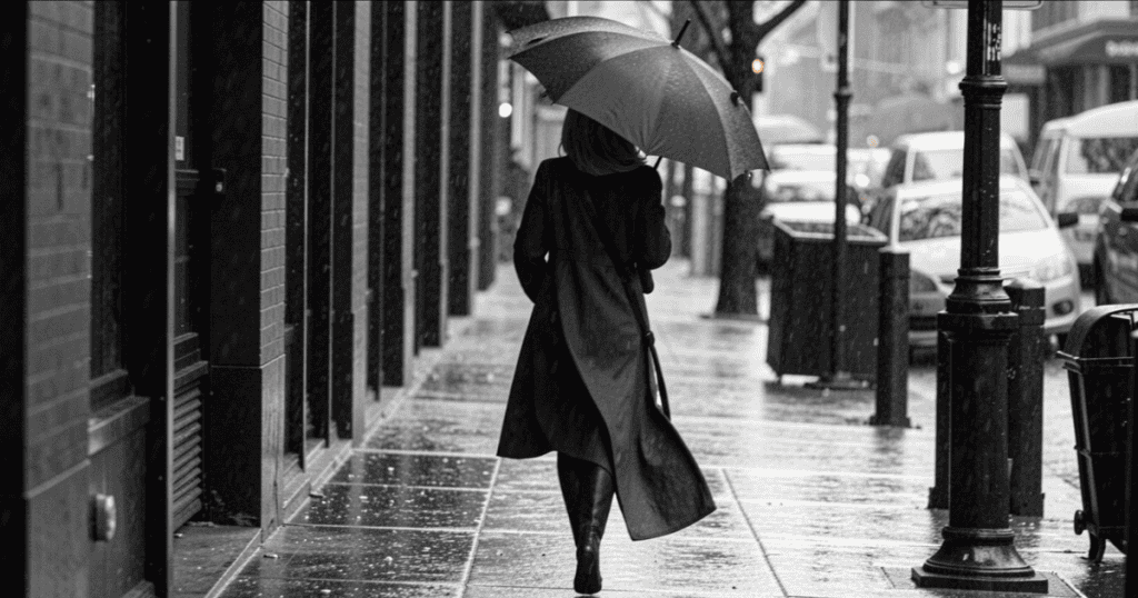 A woman in a long coat walks away from the camera down a rain-slicked city sidewalk while holding a black umbrella, moving forward with quiet resilience against a cold environment meant to restrict her.