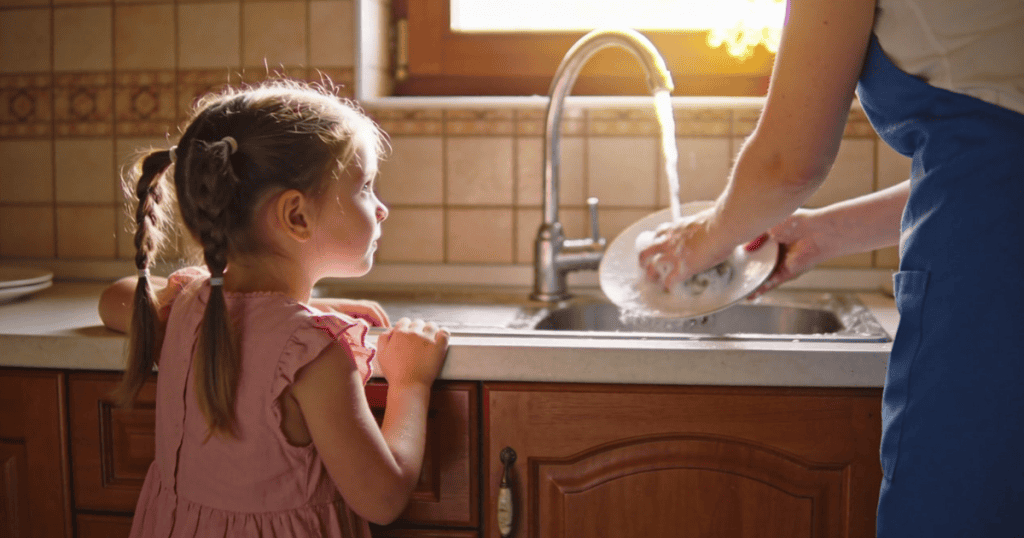 Young girl washing dishes with adult in kitchen, warm sunlight.
