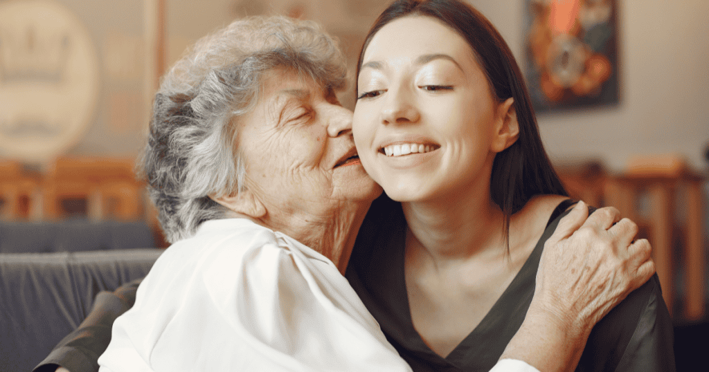 An elderly woman tenderly kissing a young woman on the cheek, representing how an enabler's warm and nurturing exterior masks the quiet harm they allow to continue in the name of love and care.