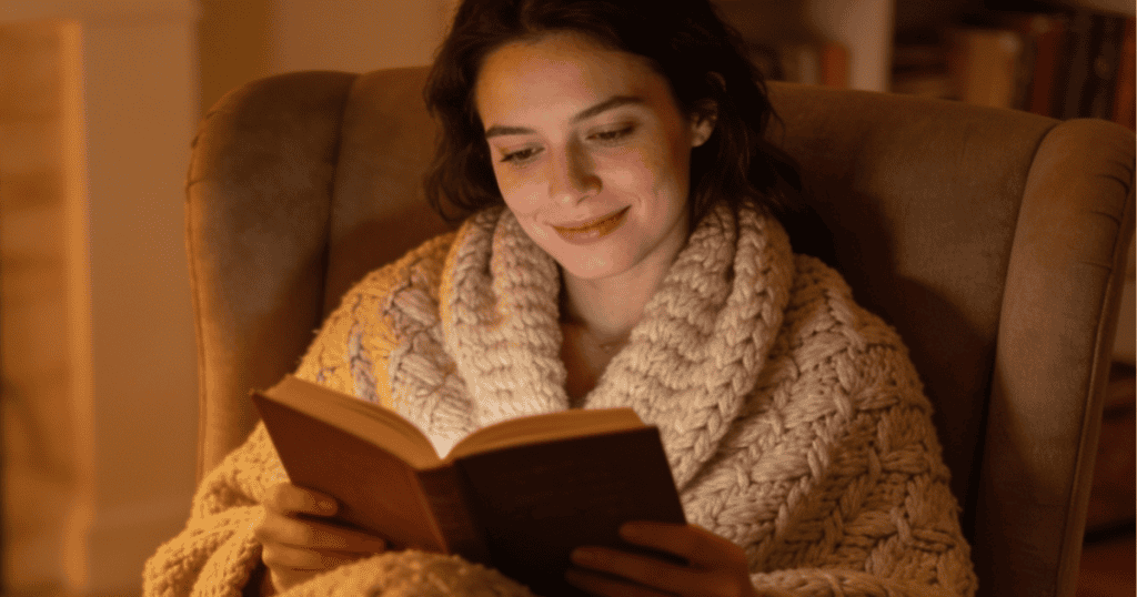 A woman sits comfortably wrapped in a cozy knit sweater while reading an open book in a softly lit room, evoking the peaceful clarity achieved once you see the enabler, you stop waiting for them to see you.