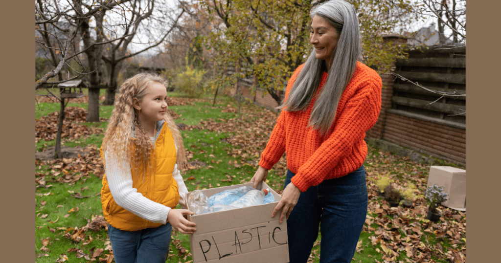 An older woman and a young girl carry a cardboard box labeled "PLASTIC" together in an autumn backyard, capturing a cooperative, nurturing routine that builds positive habits.