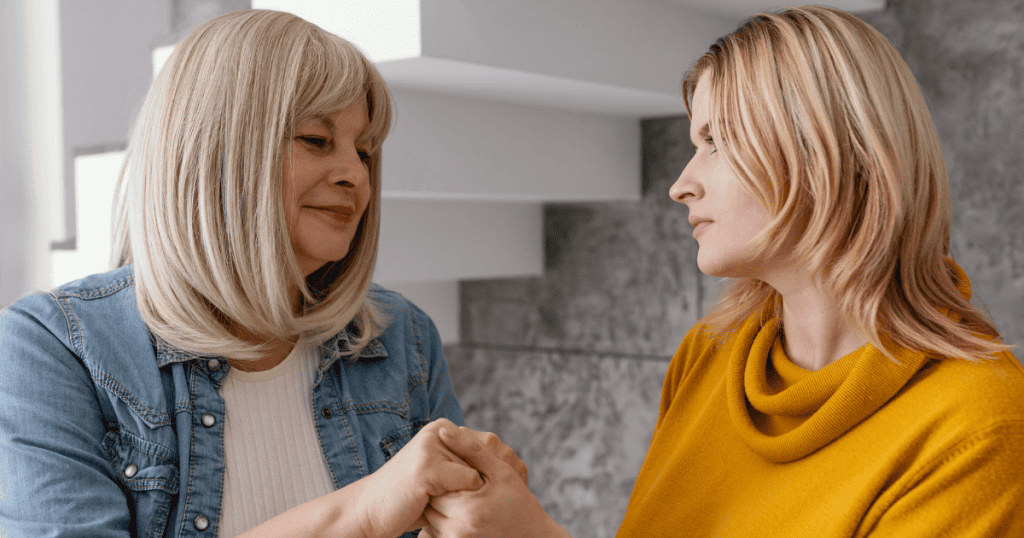 An older woman with a gentle smile holds the hands of a younger woman who looks on with quiet reservation, portraying a superficial apology used to smooth over deep-seated issues without genuine accountability.