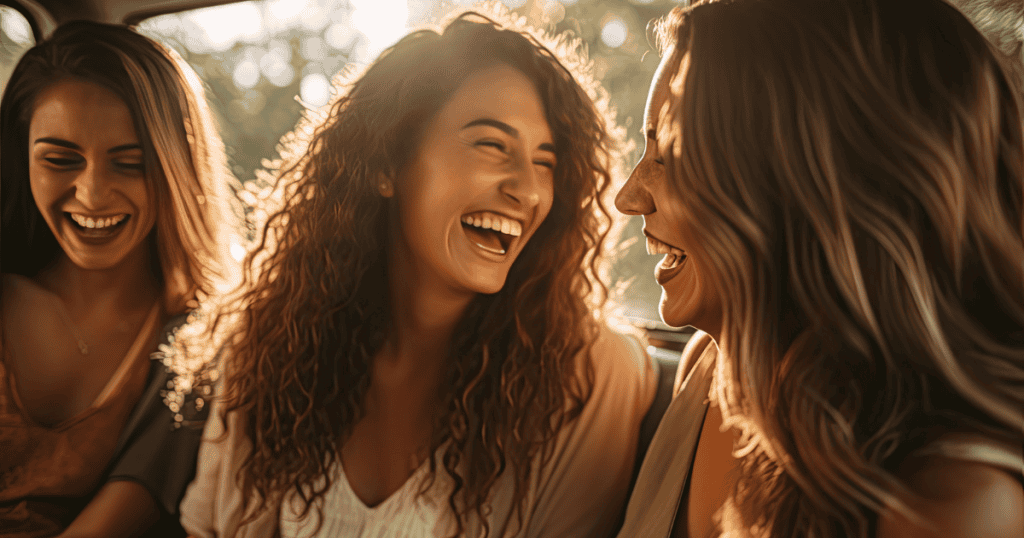 Three women share joyful, open laughter together in warm sunlight, embodying the unburdened freedom of simply living on your own terms.
