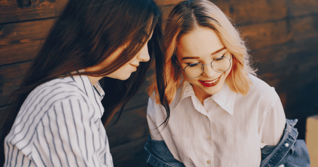 Two young women sit close together whispering and smiling over shared confidences, reflecting the vulnerability involved in placing trust in private matters.