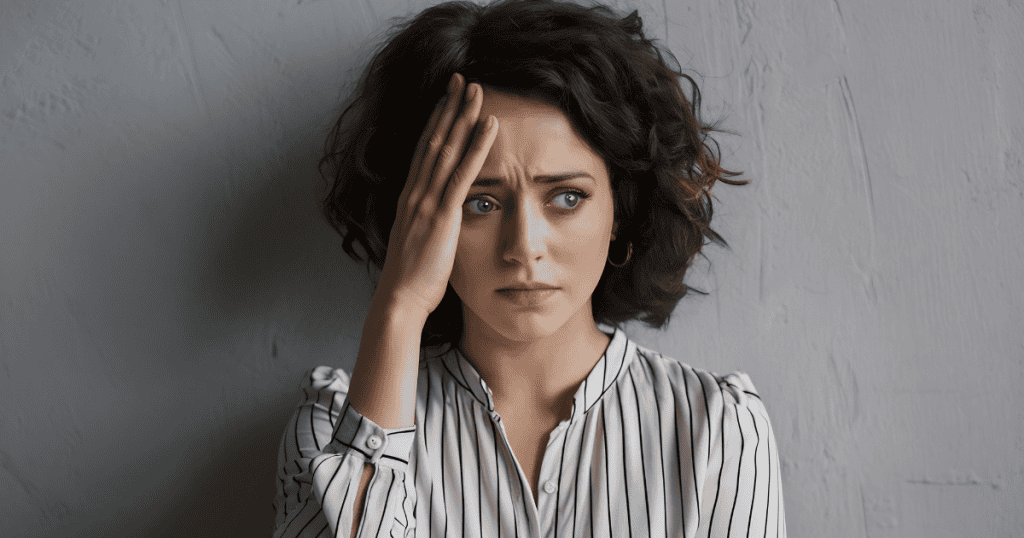 A woman with curly hair places a hand on her forehead with an anxious, distressed expression against a textured wall, portraying the overwhelming exhaustion of navigating manipulative head games.