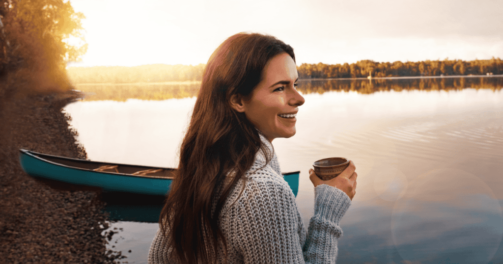 A woman smiles serenely while holding a warm mug by a calm lake with a canoe at sunset, symbolizing the grounded tranquility that remains protected from external chaos.